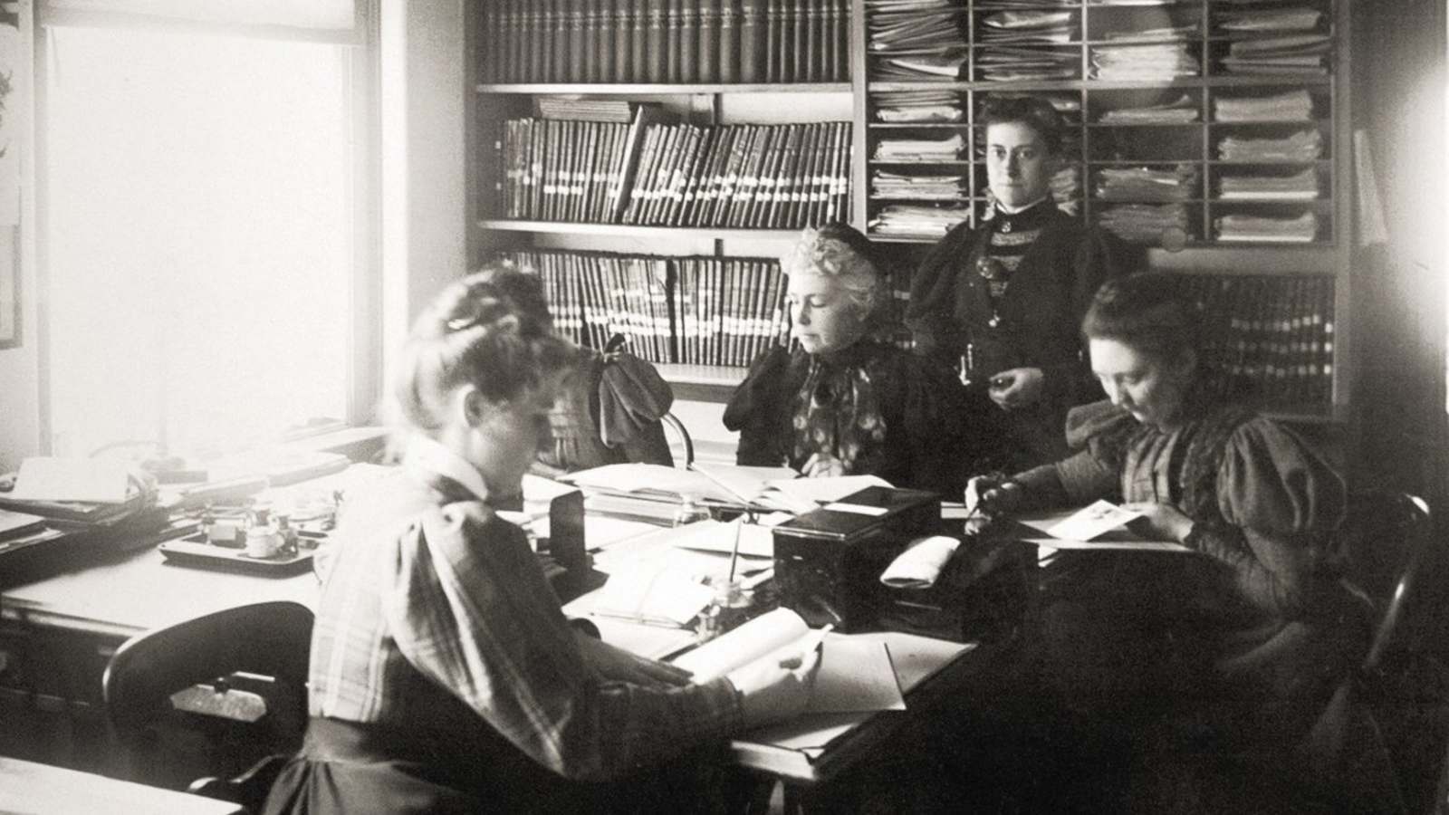 Black and white image of a group of women working at tables. Window light spills into the room. A woman with a steady gaze is standing left of center.